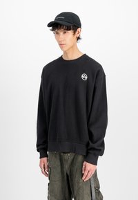 Black waffle-knit sweatshirt with ribbed cuffs and hem, featuring a circular logo on the left chest and paired with olive green cargo pants.