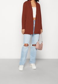 Rziavý blejzr přes bílý crop top, v kombinaci se světle modrými roztrhanými džíny a bílými teniskami. Drží malou růžovou kabelku.
