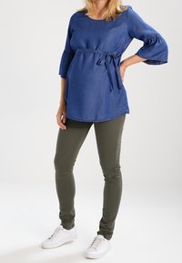 Denim tunika med midjeband i blått, med trekvartslånga klockärmar. Kombinerad med olivgröna smala byxor och vita sneakers.