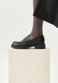 Πόδι με μαύρο χοντρό μοκασίνι με ραβδωτό πάτο, που ξεκουράζεται πάνω σε λευκό ξύλινο μπλοκ, με φεμίνιστες καλσόν σε ταυροειδές χρώμα, απέναντι από λευκό φόντο.