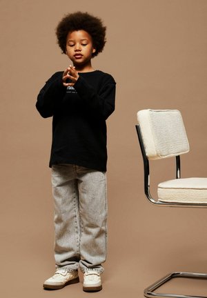 Black long-sleeve shirt with ribbed texture, gray baggy jeans, and cream sneakers. A light-colored chair is nearby against a beige background.