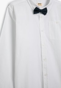 Camisa blanca de botón en tela de mezcla de algodón, con un pequeño bolígrafo en el frente y una pajarita azul marino unida al cuello.