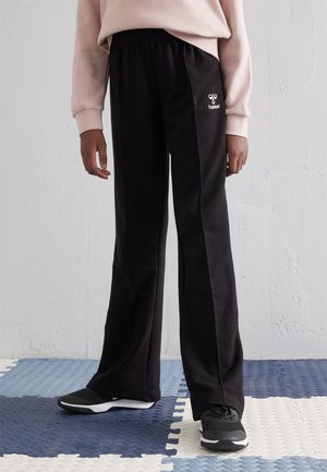 Zwarte trainingsbroek met een elastische tailleband, voorzien van een wit logo op de heup, gecombineerd met zwarte sportschoenen op een gestructureerde vloer.