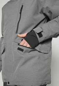 Personne portant une veste grise avec la main insérée dans une poche zippée avant et des poignets réglables avec des bandes Velcro.