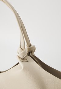 Beige skinnveske med teksturert overflate, myk form og knyttede stroppdetaljer, med en glatt indre fôr.