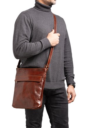 Man draagt een grijze coltrui en zwarte broek, en heeft een bruine leren schoudertas met klep en verstelbare band over zijn schouder.