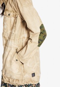 Beżowa parka z teksturowanym wykończeniem, z kamuflażowymi łatek na łokciach i ściąganym dołem. Zawiera kieszenie z przodu i metalowe akcenty.