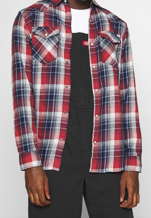 Uomo con camicia a quadri rossa, bianca e blu sopra una tuta nera e una maglietta bianca, in piedi con le braccia rilassate lungo i fianchi.