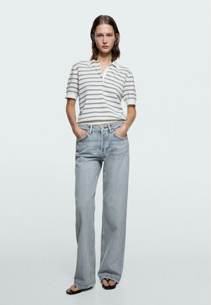 Kvinde iført en hvid og gråstribet kortærmet poloshirt, lyseblå vidde jeans og sorte sandaler, stående med hænderne i lommerne.