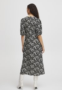 Robe noire à motifs floraux blancs, avec des manches bouffantes courtes et une coupe mi-longue. Portée avec des bottes blanches à talons.