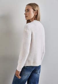 Maglione lavorato a maglia di un rosa chiaro con una finitura testurizzata, colletto e polsini a coste. Abbinato a jeans in denim blu, visto da un'angolazione.