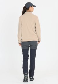 Beige fleece jack met een opstaande kraag, gecombineerd met donkergrijze en marineblauwe broeken. Het schoeisel heeft een gestructureerde zwarte zool.