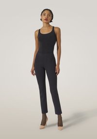 Svart bodysuiterbjuden med högmidjade svarta byxor, texturerade fishnet-strumpor och beige spetsiga klackar, mot en neutral bakgrund.