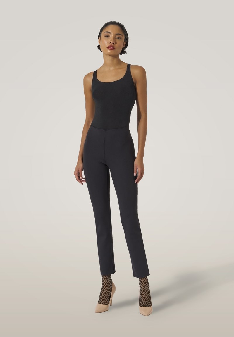 Svart bodysuiterbjuden med högmidjade svarta byxor, texturerade fishnet-strumpor och beige spetsiga klackar, mot en neutral bakgrund.