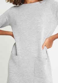 Donna che indossa un vestito a maglia grigio chiaro a maniche corte con due tasche anteriori, mani appoggiate dentro le tasche.