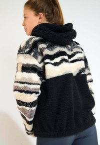 Giacca in pile con un pattern a onde nero e crema, superficie testurizzata e cappuccio. Orlo e polsini elastici visibili.