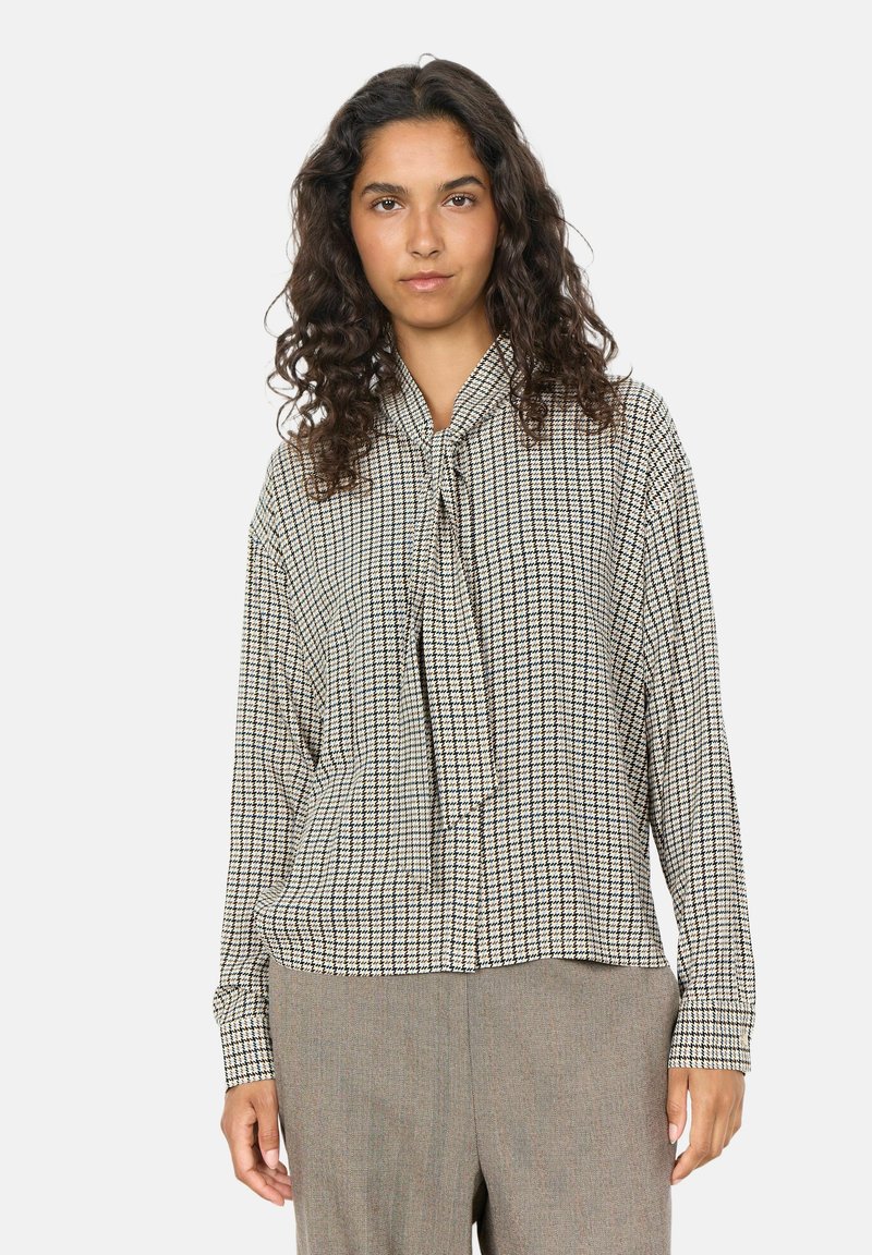 Femme aux cheveux bouclés et foncés porte un chemisier à manches longues à carreaux avec un nœud devant et un pantalon marron clair, debout devant un fond uni.