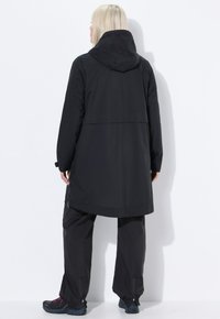 Persona con capelli biondi che indossa un lungo giubbotto nero con cappuccio, pantaloni neri e scarpe da trekking scure, in piedi di fronte a un muro bianco.