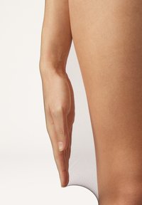 Durchsichtige tanfarbene Strümpfe bis zum Oberschenkel, mit glatter Textur und zartem, durchscheinendem Finish, die an einem gebräunten Bein von einer Hand angelehnt sind.