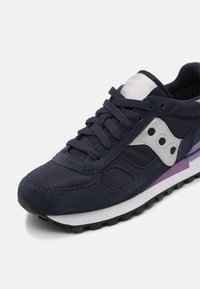 Chaussure de course bleu marine avec logo blanc, dessus en mesh et suède, lacets noirs et semelle blanche avec accents violets sur un fond clair.