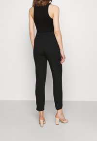 Svarta, smala byxor med en slät yta och elastisk midja. Matchas med ljusrosa blockklack-sandaler. Vy bakifrån.