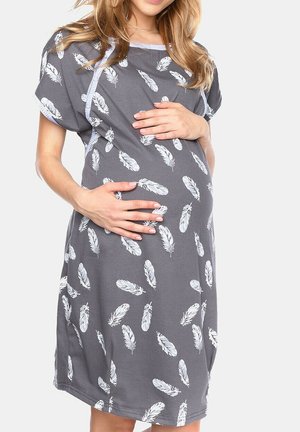 Pregnant woman in a gray dress with white feather patterns, gently holding her belly with both hands against a plain background.