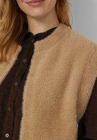 Beige faux bontvest met een ronde halslijn, gedragen over een donkerbruine blouse met knopen. Zachte textuur, subtiele glans en contrasterende kleuren.