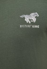 Dunkelgrünes T-Shirt mit einem grafischen Motiv eines weißen Pferdes und dem Text "MUSTANG JEANS" darunter. Der Stoff wirkt glatt und das Design ist schlicht.