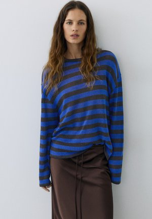 Femme aux longs cheveux ondulés portant un pull rayé bleu et noir et une jupe marron, debout contre un mur blanc.