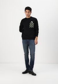 Sort sweatshirt med tekstureret grafisk lomme i grønne nuancer. Slim-fit jeans og sorte ankelstøvler. Simpelt, minimalistisk design.