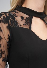 Vestido negro con hombros de encaje transparente con patrón floral, cuello alto y detalle de abertura en la parte delantera. Textura suave con cuerpo ajustado y liso.