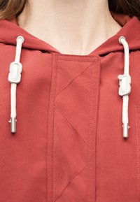 Κοντινό πλάνο ενός ατόμου που φοράει φούτερ με κουκούλα σκουριάς, με λευκά κορδόνια και ασημένιες μεταλλικές άκρες κοντά στην περιοχή του λαιμού.