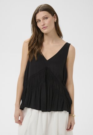 Žena u crnoj majici bez rukava s V-izrezom i teksturiranim dijagonalnim uzorcima te bijeloj suknji na jednobojnoj pozadini.