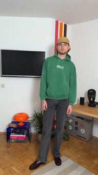 Πράσινο φούτερ με κουκούλα και μπροστινή τσέπη με λευκό λογότυπο, συνδυασμένο με γκρι καμπάνα τζιν και μαύρα λαμπερά παπούτσια. Το φόντο έχει πολύχρωμο τοίχο και διακόσμηση.