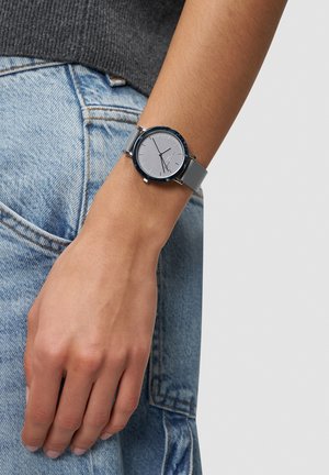 Ronde zilveren horloge met een grijze lederen band. De wijzerplaat heeft minimalistische markeringen en een glad zilveren wijzerblad met een glanzende rand.
