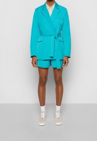 Blazer turchese con cintura a cravatta e tasche, abbinato a pantaloni corti coordinati. Camicia bianca con motivo floreale sotto e sneakers chiare.