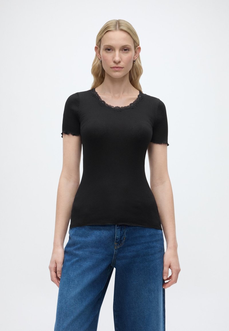 Ξανθιά γυναίκα που φοράει στενό μαύρο ριμπ με κοντά μανίκια και δαντελένια λεπτομέρεια, μαζί με χαλαρά μπλε τζιν, στέκεται μπροστά σε ένα απλό φωτεινό φόντο.