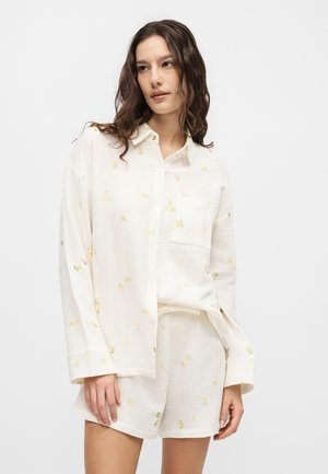 Vrouw met lang golvend haar, gekleed in een wit overhemd met knopen en bijpassende korte broek met kleine gele bloemenprints, staand voor een effen achtergrond.