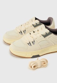 Zapatos deportivos beige con paneles de malla, suelas de goma gruesas y cordones color crema. Incluye un par de cordones crema adicionales junto a los zapatos.