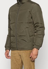 Vadderad olivgrön jacka med blixtlås, hög krage, sidofickor och elastiska manschetter. Slät textur med stickade paneler.