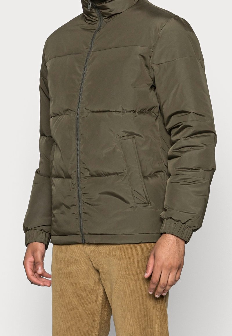 Vadderad olivgrön jacka med blixtlås, hög krage, sidofickor och elastiska manschetter. Slät textur med stickade paneler.