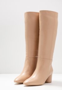 Beige leather knee-high boots with a smooth texture, pointed toes, and block heels. Minimalistic design with no visible hardware or patterns.