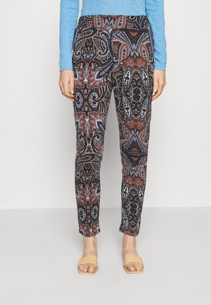 Kobieta ubrana w dopasowane spodnie z wysokim stanem ozdobione brązowym, czarnym i niebieskim wzorem paisley, w połączeniu z niebieską bluzką z długim rękawem i beżowymi sandałami z odkrytymi palcami.