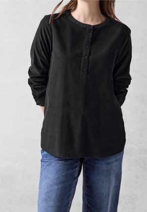 Frau trägt ein langärmeliges schwarzes Hemd mit Knöpfen und blaue Jeans, steht mit den Händen hinter dem Rücken vor einem einfachen Hintergrund.