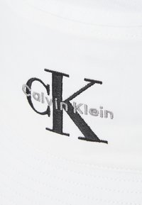 Balta medvilninė skrybėlė su plačiu kraštu, kuriame išsiskiria juodas "CK" logotipas ir pilkos siuvinėjimo raidės "calvin klein" priekyje.