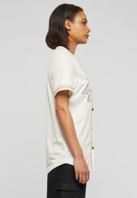 Maglietta da baseball crema con maniche corte, caratterizzata da strisce beige a contrasto sulle maniche e una chiusura frontale con bottoni. Tessuto liscio, vestibilità comoda.