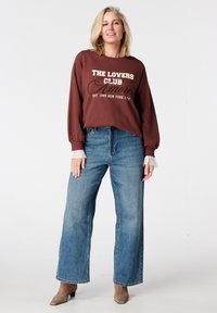 Bruine trui met de tekst "THE LOVERS CLUB", gecombineerd met blauwe, high-waisted jeans met wijde pijpen. Crèmekleurige kantaccenten bij de manchetten. Beige enkellaarzen.