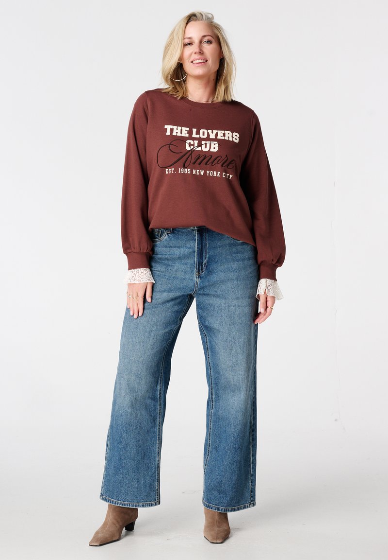 Bruine trui met de tekst "THE LOVERS CLUB", gecombineerd met blauwe, high-waisted jeans met wijde pijpen. Crèmekleurige kantaccenten bij de manchetten. Beige enkellaarzen.