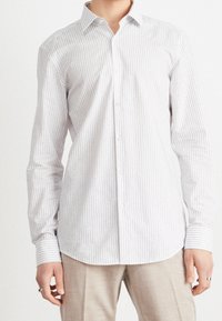 Man in een witte langarmige overhemd met dunne zwarte verticale strepen en beige broek, staande tegen een witte achtergrond.
