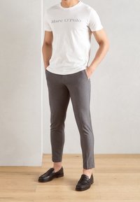 Bílá tričko s textem "Marc O'Polo", šedé zúžené kalhoty, černé kožené mokasíny s perforovanými detaily, na světlém pozadí.
